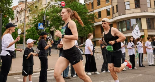 Female runner in black athletic wear leads a street race, with a male runner beside her and spectators along a city sidewalk behind them.
