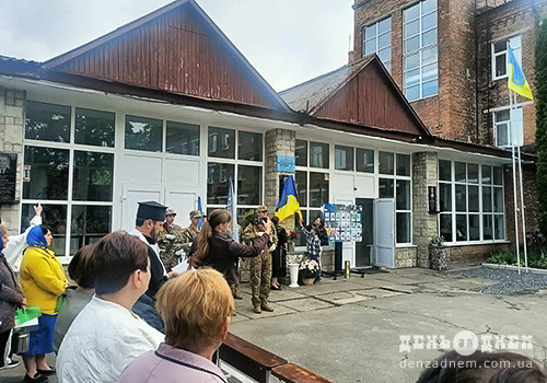 Group of people gathered outside a school-like building for a ceremony, with a Ukrainian flag and soldiers in uniform visible.