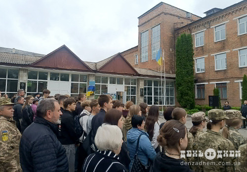 Crowd of students and adults gathered outside a brick school building, with soldiers in camouflage and a Ukrainian flag on a flagpole.