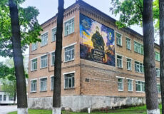 Three-story brick building framed by trees, with a large mural of a soldier on the corner wall.