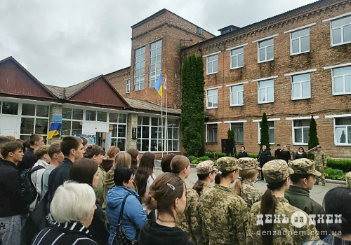 Crowd of students and cadets outside a brick school listening to speakers during a flag-raising ceremony with a Ukrainian flag visible outside the building.