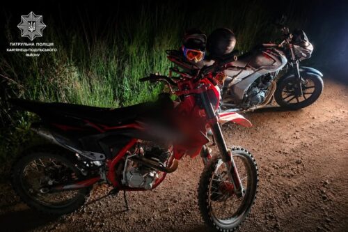 Two off‑road motorcycles parked on a dim dirt road at night, riders wearing helmets nearby, police insignia visible in the upper left.