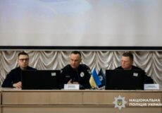 Three Ukrainian police officials sit at a panel desk on a stage during a press conference; laptops are open, with a small Ukrainian flag and police insignia on the table.