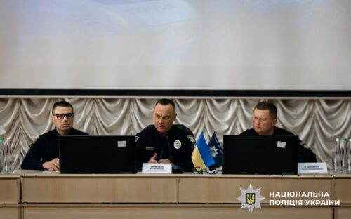 Three Ukrainian police officials sit at a panel desk on a stage during a press conference; laptops are open, with a small Ukrainian flag and police insignia on the table.