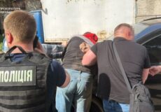 Ukrainian police officer in a vest labeled поліція arresting a man beside a dark car.