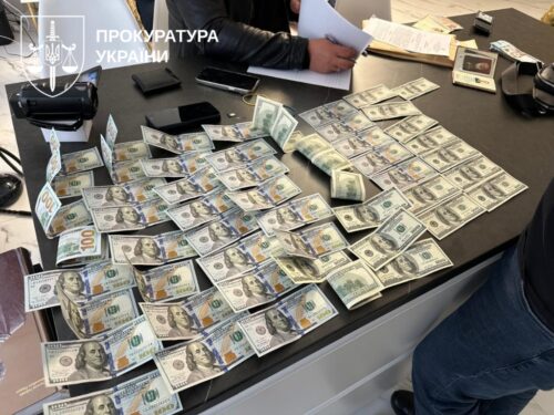 Cash-strewn office desk with bundles of U.S. dollars, a person writing in the background, and a Ukrainian Prosecutor's Office watermark in the corner.