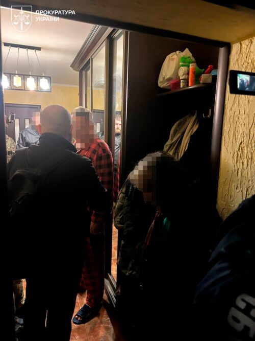 Group of people with blurred faces in a dim residential hallway; open wardrobe with clothes and bags, a video camera records the scene, Ukrainian prosecutor's office watermark in corner.