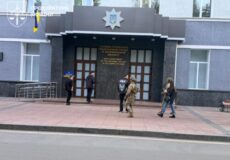 Front entrance of a government building displaying a large emblem and Ukrainian flag, with four people walking on the sidewalk in front.