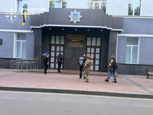 Front entrance of a government building displaying a large emblem and Ukrainian flag, with four people walking on the sidewalk in front.