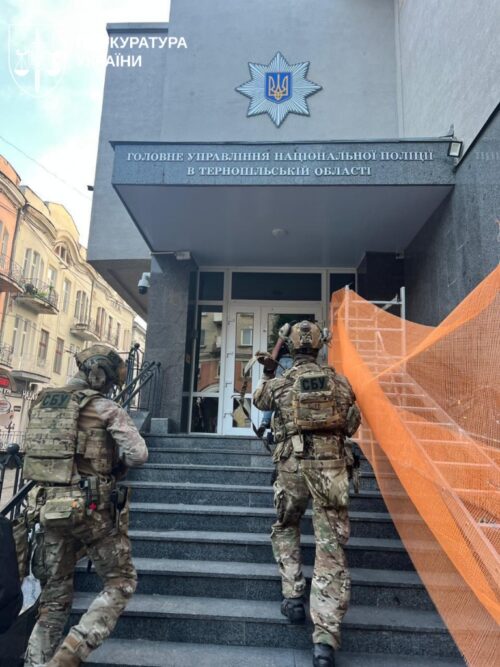 Two armed soldiers in camouflage walk up the steps toward a Ukrainian police building entrance with a sign for the National Police of Ukraine visible above the door.