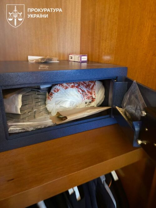 Open blue safe on a wooden shelf, filled with stacked envelopes and plastic bags; Ukrainian Prosecution logo in the top-left corner.