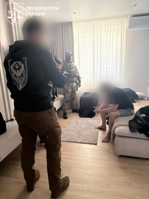 Armed officers from Ukraine's Prosecutor's Office confront a seated man in a living room, with another officer in camouflage near the window; logo visible in the top-left corner.