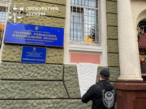 Facade of Ukraine's Prosecutor's Office with a blue National Police sign; a man in a black jacket with a police emblem stands beside a column outside the building.