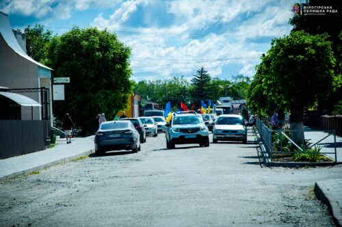 Cars driving down a sunlit street in a small town, with colorful flags lining the road for a parade visible in the distance.