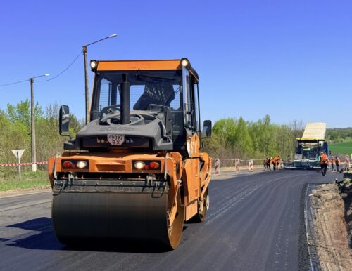 Orange road roller smoothing fresh asphalt on a rural road; workers and a paving machine visible in the distance