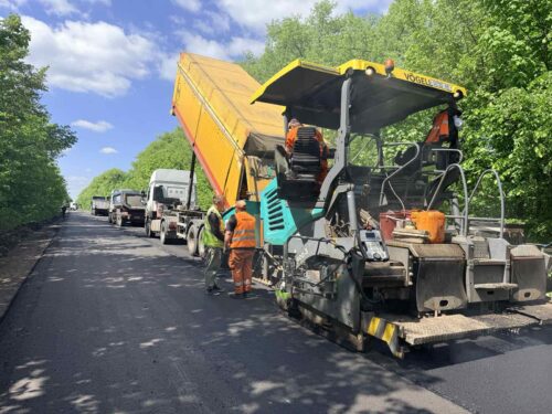 Road construction scene: an asphalt paver lays fresh pavement while an orange dump truck is raised; workers in orange vests stand nearby with vehicles queued behind.