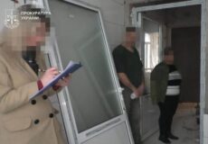 A woman in a beige coat writes on a clipboard beside a doorway in an unfinished interior, with a Ukrainian Prosecutor's Office watermark in the corner.