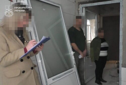 A woman in a beige coat writes on a clipboard beside a doorway in an unfinished interior, with a Ukrainian Prosecutor's Office watermark in the corner.