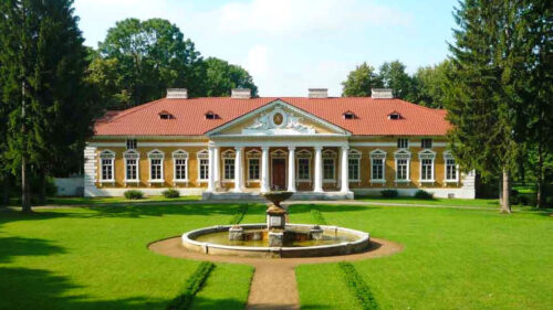 Yellow neoclassical villa with a white columned portico and red tiled roof, set in a manicured park with a circular fountain in front.