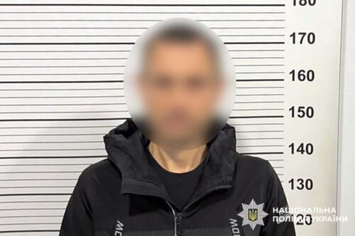 Blurred man in a dark jacket stands against a height chart in a police mugshot setup.