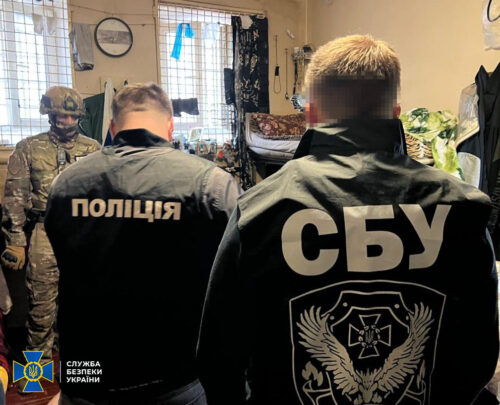 Two men with back jackets reading 'ПОЛІЦІЯ' and 'СБУ' face away, a soldier in camouflage stands in a cluttered room.
