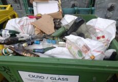 Green glass recycling bin overflowing with bottles, bags, and paper/cardboard waste.