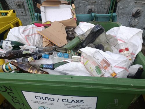 Green glass recycling bin overflowing with bottles, bags, and paper/cardboard waste.