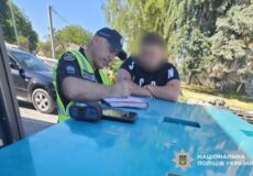 Ukrainian police officer in a reflective vest signs a document on a blue police car as a blurred civilian watches nearby.
