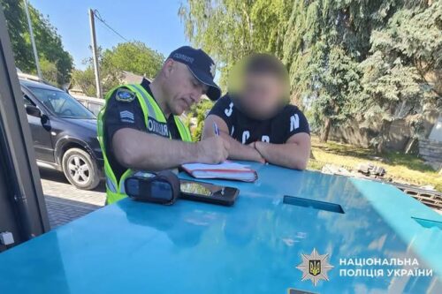 Ukrainian police officer in a reflective vest signs a document on a blue police car as a blurred civilian watches nearby.