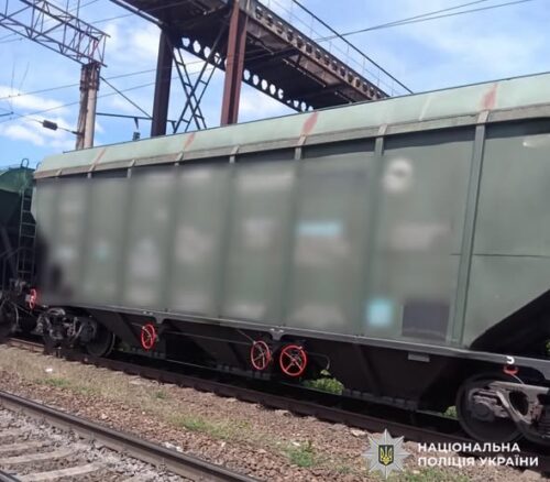 Green freight wagon on railway tracks beside a metal bridge, with a Ukrainian National Police watermark in the corner.
