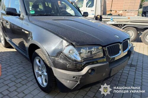 Black BMW SUV with visible front-end damage (scuffed bumper and fender) parked on a cobblestone lot; Ukrainian National Police watermark in corner.