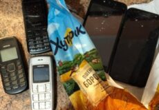 Cluster of old mobile phones and two smartphones scattered on a countertop beside a torn snack bag.