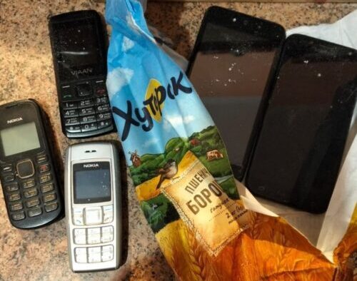 Cluster of old mobile phones and two smartphones scattered on a countertop beside a torn snack bag.