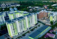 Aerial view of a modern lime-green and pale-yellow apartment complex with a construction site nearby and a Ukrainian Prosecutor's Office logo in the corner.