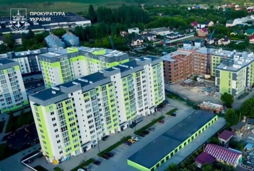 Aerial view of a modern lime-green and pale-yellow apartment complex with a construction site nearby and a Ukrainian Prosecutor's Office logo in the corner.