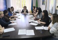 Group of ten women seated around a conference table in a meeting room, papers and notebooks laid out in front of them.