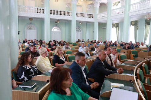 Large conference hall filled with attendees seated in green chairs, listening to a presentation in a pale green room with columns and balconies.