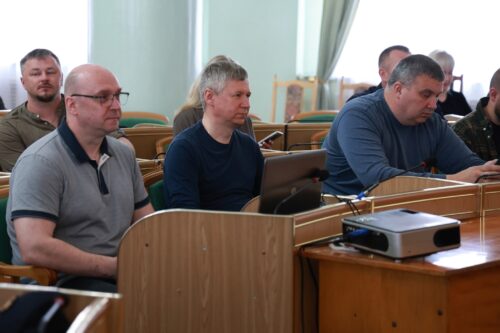 Men seated in a conference room listening to a presentation; a projector sits on the front desk.