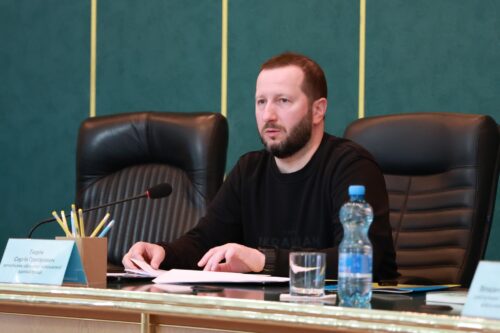 Man with a beard sits at a conference table with a microphone, papers, pencils, a water bottle, and a glass in front of him, appearing to speak or listen during a formal meeting.