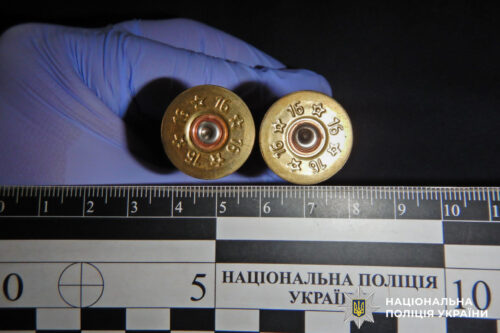 Two spent brass cartridge cases held by a gloved hand above a measurement ruler, with a Ukrainian police badge in the corner.