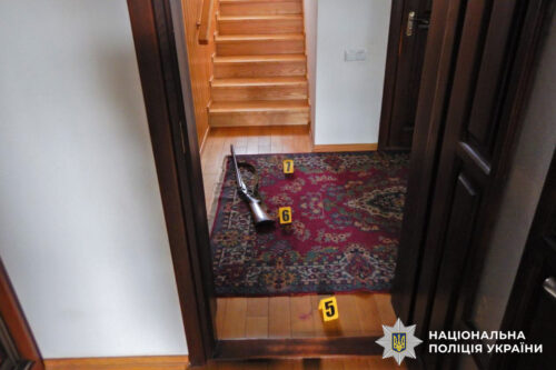 Hallway crime scene with a rifle on a patterned rug and yellow evidence markers 5 and 7; stairs ahead, police watermark visible.