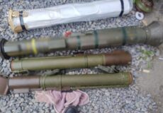 Several cylindrical munitions laid out on gravel, likely rocket launch tubes; a pink cloth and a shoe sit nearby; Ukrainian prosecutor watermark in the corner.