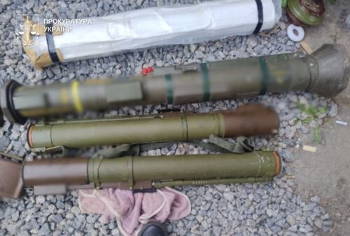 Several cylindrical munitions laid out on gravel, likely rocket launch tubes; a pink cloth and a shoe sit nearby; Ukrainian prosecutor watermark in the corner.