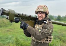Soldier in camouflage gear carrying a shoulder-fired rocket launcher across a grassy field, outdoors under a cloudy sky.