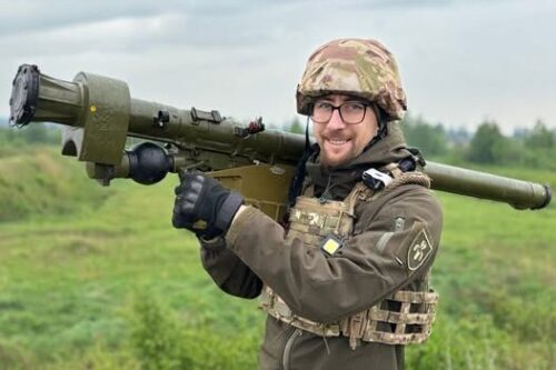 Soldier in camouflage gear carrying a shoulder-fired rocket launcher across a grassy field, outdoors under a cloudy sky.