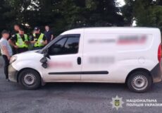 White panel van stopped on a road; four police officers in high-visibility vests speak with a man; Ukrainian National Police logo appears bottom-right.