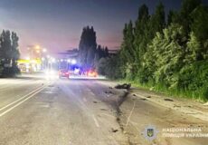 Nighttime road collision with debris scattered across lanes, a damaged car, and flashing emergency lights in the distance, trees on the right side.