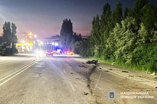 Nighttime road collision with debris scattered across lanes, a damaged car, and flashing emergency lights in the distance, trees on the right side.