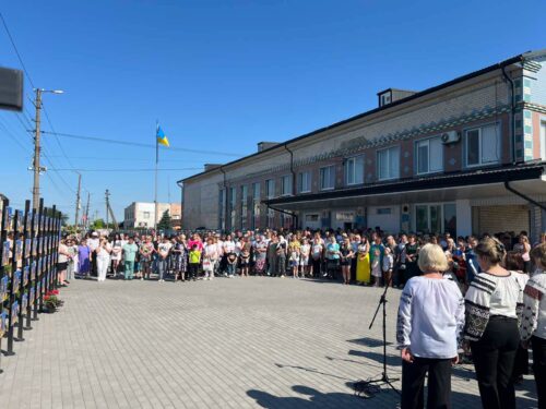 Crowd of people gathered outside a brick building on a sunny day, with a Ukrainian flag fluttering nearby.