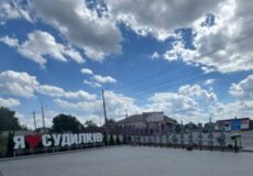 Outdoor plaza with a large sign reading 'Я люблю СУДИЛКІВ' and a row of blue decorative bikes along a fence under a cloudy blue sky.